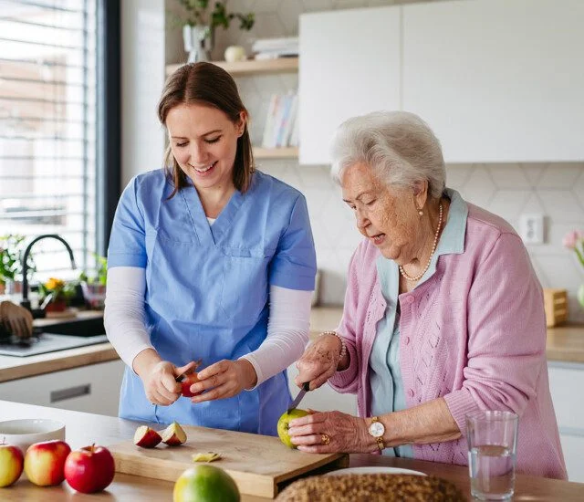 Home caregiver and elderly woman baking apple pie together, cutting apples. Household chores for senior woman, help from nurse.