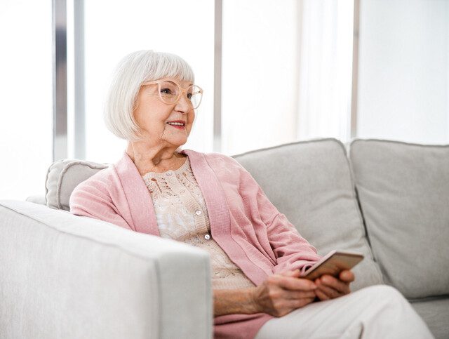 Senior woman dressed in pink sitting on gray couch with smartphone in hand.