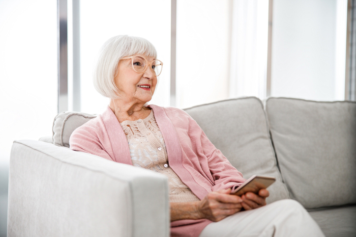 Senior woman dressed in pink sitting on gray couch with smartphone in hand.