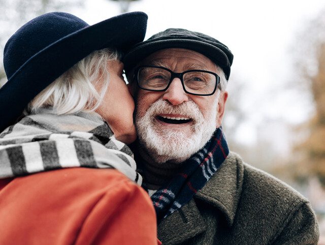 Senior woman kissing a smiling senior man on the cheek outdoors.