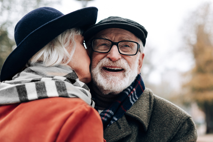 Senior woman kissing a smiling senior man on the cheek outdoors.